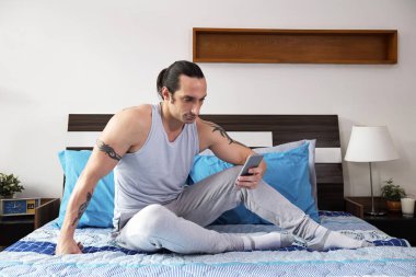 Elindeki yatağa oturma ve yatak odasında onun smartphone cep telefonu ile mesaj okuma dövmeli genç yakışıklı adam