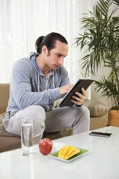 年轻的严肃男子使用平板电脑 而在沙发上休息与新鲜水果和水在桌子上的玻璃 — 图库照片