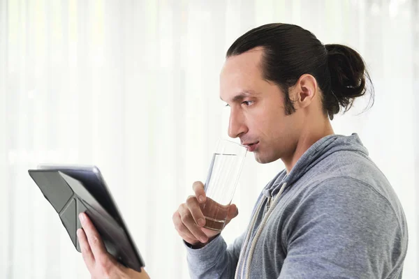 Stilig Ung Man Dricksvatten Medan Läser Något Digitala Tablett — Stockfoto