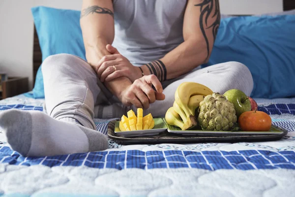座っていると 彼の寝室で熟した果物のトレイが付いているベッドで休んで肩の入れ墨をした男をクローズ アップ — ストック写真
