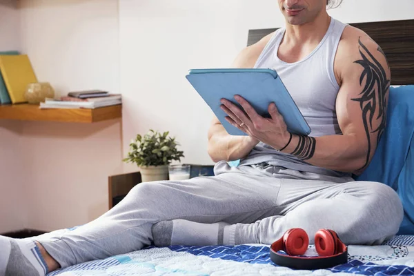 ベッドで休むと家でデジタル タブレットを使用してオンラインの通信手の入れ墨をした若い筋肉男 — ストック写真