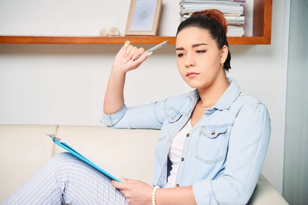 Pensive Young Woman Sitting Couch Pen Clipboard Her Knees Making — Stock Photo, Image