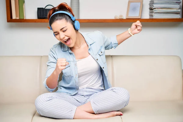 Young pretty girl in casual clothing sitting on couch in headphones and singing
