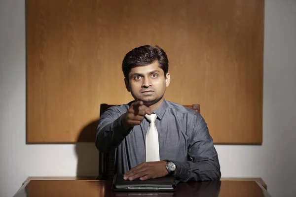 Displeased Indian entrepreneur sitting at table and pointing at camera