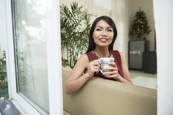 微笑的亚洲女人在沙发上享受她的闲暇时间 喝着咖啡 一边做梦 一边望着敞开的窗户 — 图库照片
