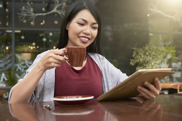 Hymyilevä Aasialainen Nainen Juo Kahvia Käyttää Hänen Digitaalinen Tabletti Istuessaan — kuvapankkivalokuva