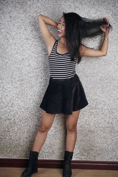 Happy Asian woman in skirt and dark long hair standing near the wall and posing