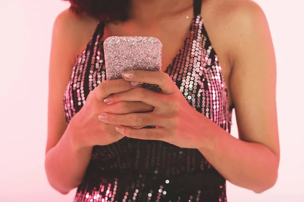 Crop view of woman in black top with silver sequins holding in hands mobile phone covered with silver glitter over pink studio background