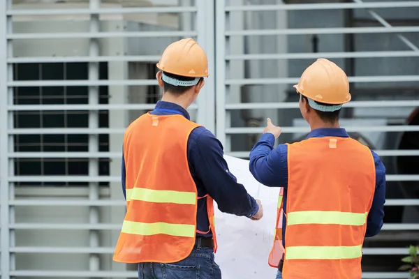 站在建筑工地上的男子 穿着安全帽和背心 拿着草稿指着建筑物 — 图库照片