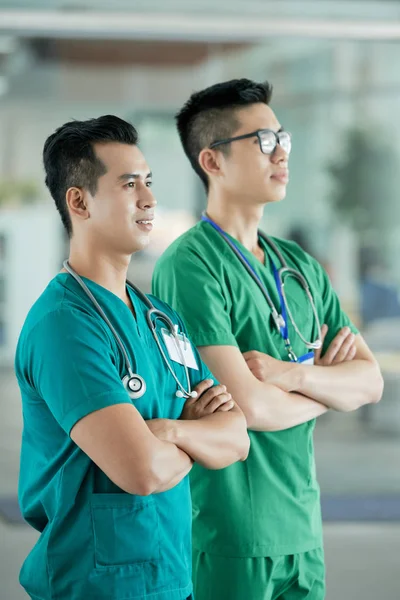 Two positive Asian men in medical scrubs keeping arms crossed and looking away while standing on blurred background of hospital hall