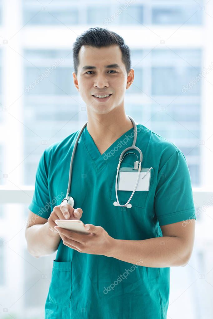 Handsome Asian guy in medical uniform smiling and looking at camera while standing in hospital and browsing smartphone