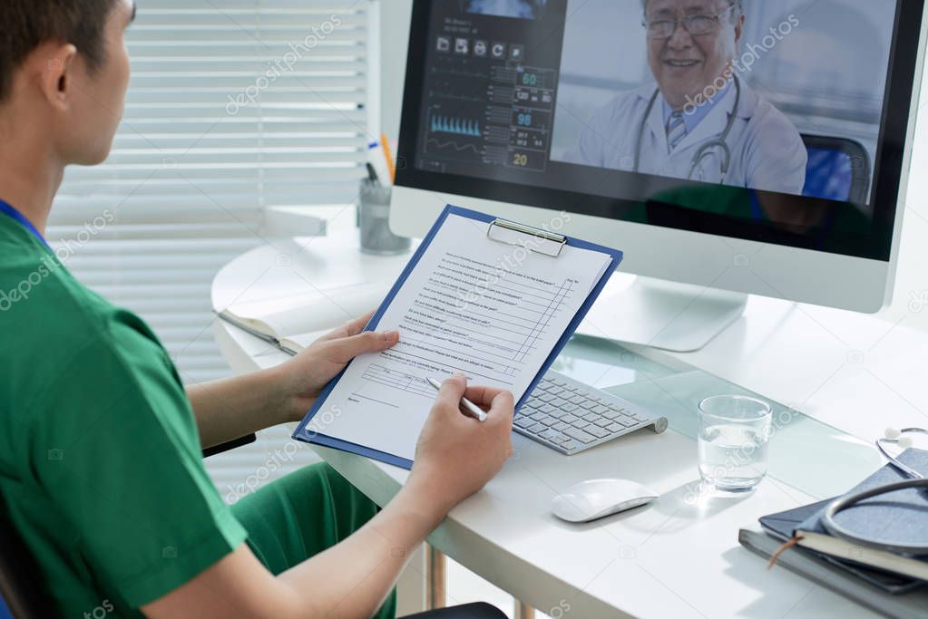 Unrecognizable Asian health professional with clipboard sitting at table in hospital office and taking part in video conference