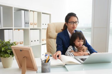 Masada ofiste diz küçük kızla ciddi bilgisayar kullanırken çalışan yetişkin Asyalı kadın