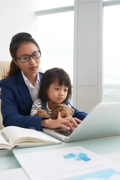 アジアの女性のスーツと眼鏡のオフィスで膝の上に座って魅力的な少女と作業テーブルでノート パソコンを使用しての — ストック写真