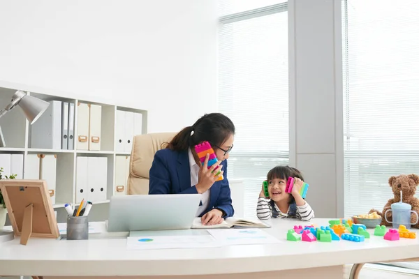 成人亚洲正式女商人与顽皮的小女孩坐在桌子上在现代办公室与五颜六色的块的乐趣 — 图库照片