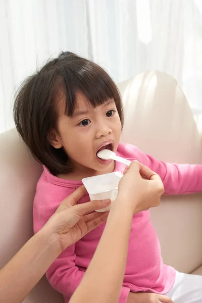 在阳光下坐在沙发上的亚洲小女孩用牛奶酸奶给亚洲小女孩的手 — 图库照片