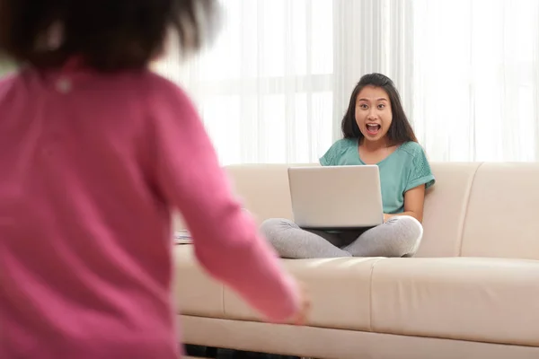 亚洲情绪激动的女人坐在沙发上 拿着笔记本电脑 看到跑小女儿 显得很兴奋 — 图库照片