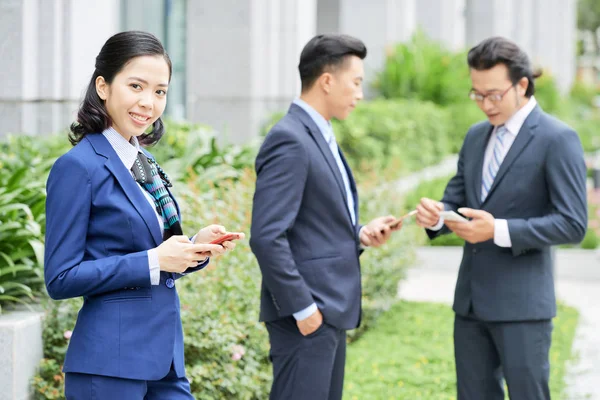 スマート フォンの背景に男性の同僚と屋外で立ってと陽気なエレガントなアジアの女性 — ストック写真