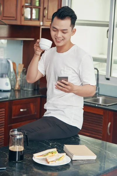 朝のコーヒーを飲んで 笑顔で電話を使用して キッチンのカウンターの上に座ってカジュアルなアジア男 — ストック写真