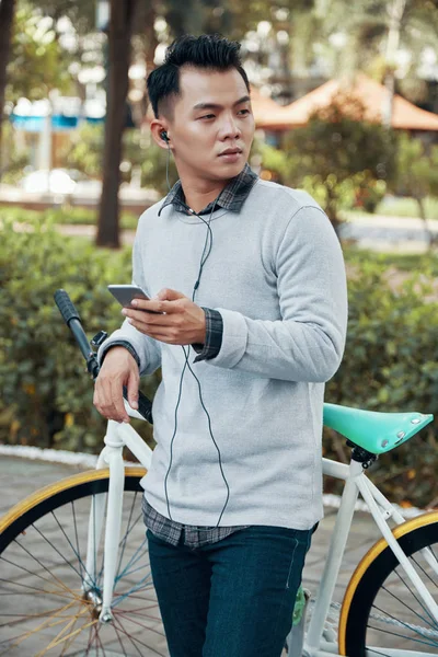 잘생긴 아시아 스마트폰 자전거에 공원의 흐리게에 이어폰에서 음악을 즐기는 — 스톡 사진