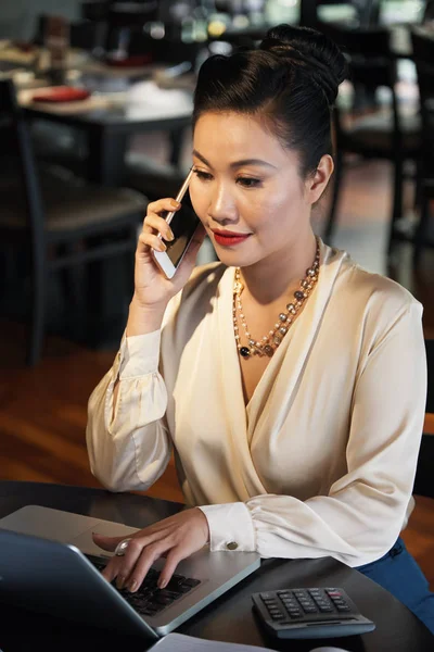 Asian woman in white elegant blouse and necklace sitting at wooden table talking on mobile phone