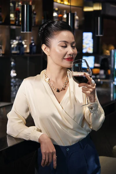 Gorgeous young Asian delighted woman standing relaxed leaning on dark bar counter and smelling red wine in glass with closed eyes