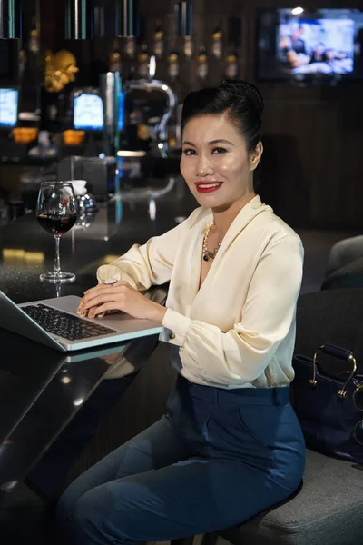 华丽愉快的微笑的亚洲女人在优雅的衣服坐在酒吧柜台与一杯葡萄酒和笔记本电脑 — 图库照片