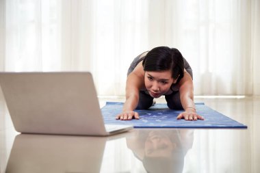 Beautiful Chinese woman exercising yoga while watching instructional videos on laptop at home  clipart
