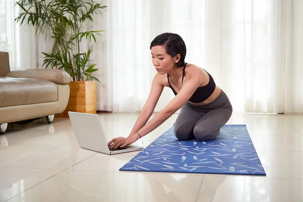 アジアン フィット若い女性運動マットの上の床に座って オンライン スポーツ トレーニング自宅を探して彼女のラップトップで入力 — ストック写真