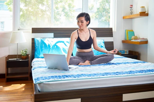 ベッドの使い方が彼女のラップトップ コンピューターと彼女の寝室で瞑想になるの蓮華座に座っているアジアのきれいな女性 — ストック写真