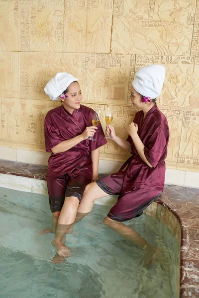 Young Asian women in gowns and towels on their heads drinking champagne and talking to each other in swimming pool