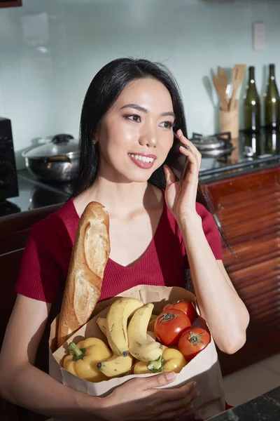 食料品の袋が付いている台所に立っていると電話で話している若い笑顔のベトナム人女性 — ストック写真