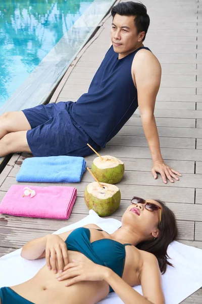 年轻的美丽的女人戴着太阳镜躺在池畔晒太阳 和她的男朋友坐在她身边 喝着椰子鸡尾酒 — 图库照片