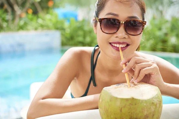 Portrait of happy young girl in sunglasses drinking coconut drink from straw while sunbathing outdoors in summer day