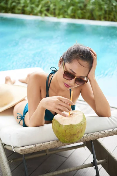 年轻的美丽的女人戴着太阳镜躺在游泳池边附近的日光浴床上 在户外喝椰子鸡尾酒 — 图库照片
