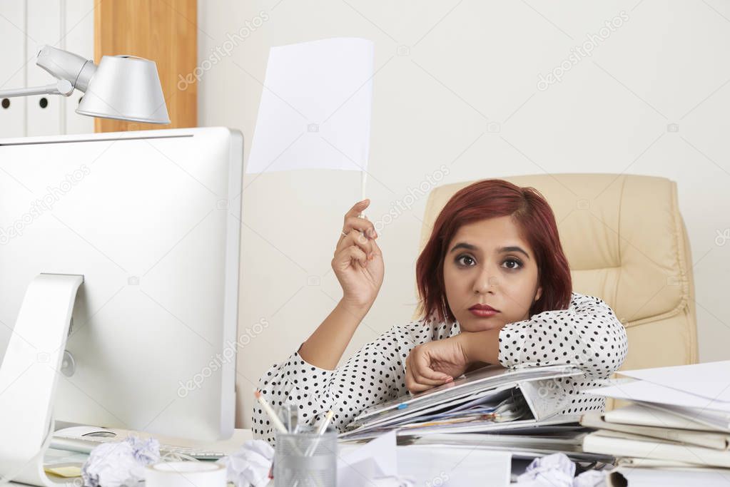 Exhausted young businesswoman sitting at office table with heap of documents and holding white flag