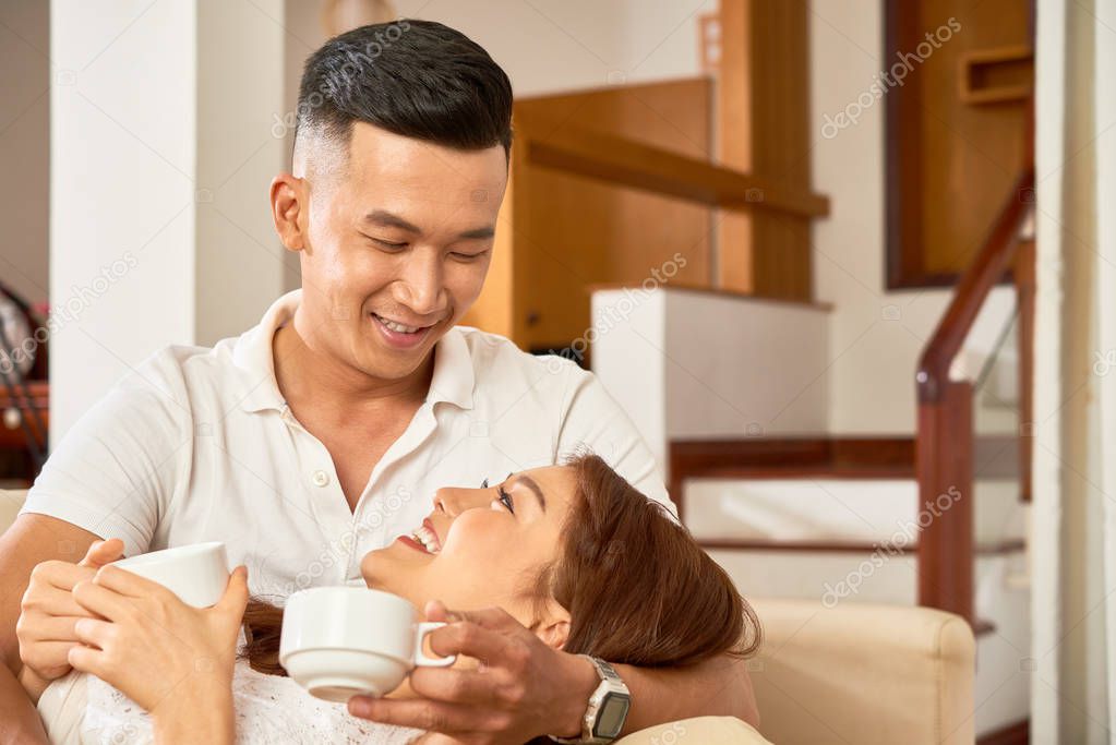 Smiling Asian couple embracing and drinking coffee together while relaxing on sofa at home