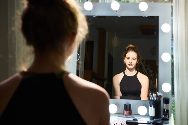 Молодая красивая модель сидит и смотрит в зеркало, она восхищается своей красотой и готова к работе
