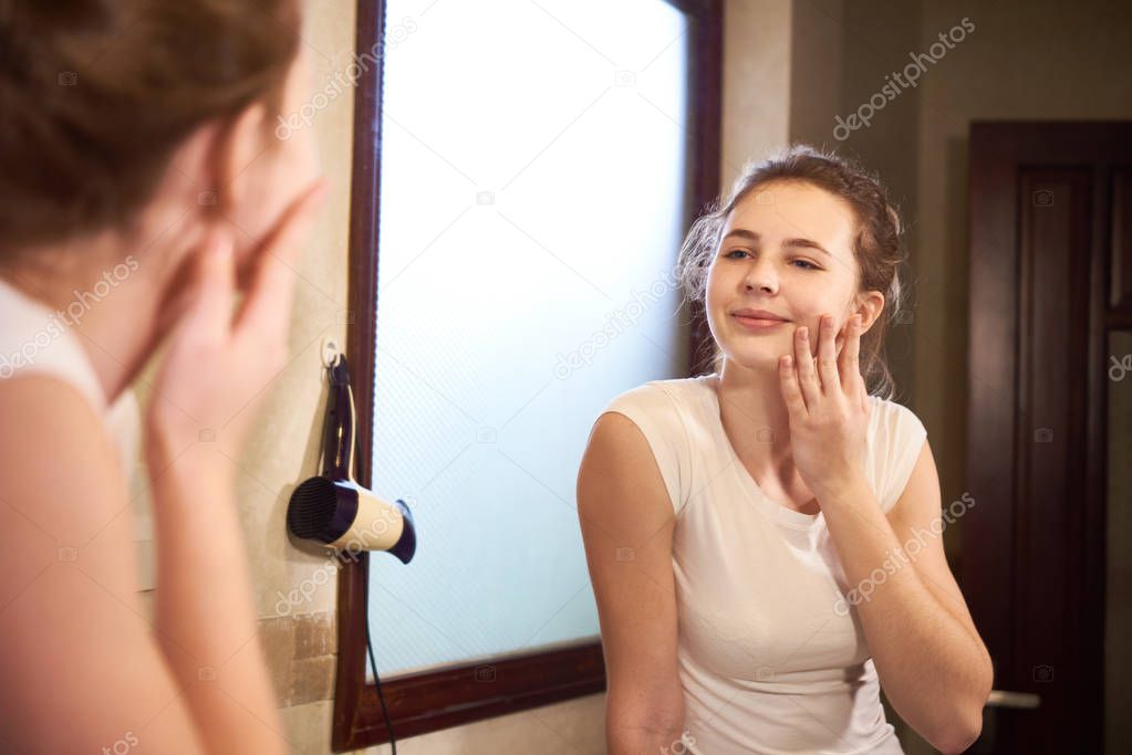 Young beautiful woman touching her face while looking at mirror in bathroom