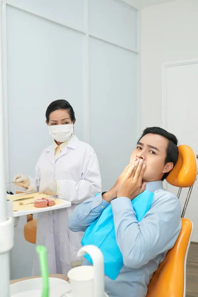 Adult Asian man covering mouth in fear while sitting in dental char in office with doctor