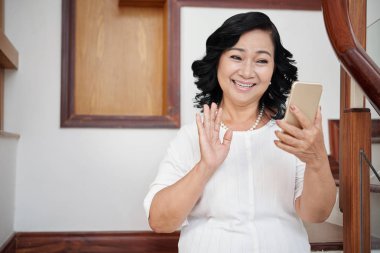 Akrabaları ile smartphone ile görüntülü sohbet sahip, el sallayarak ve neşeyle gülümseyen çekici üst düzey Asyalı kadın