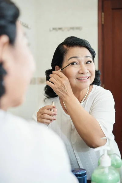迷人的亚洲老年妇女的肖像与珍珠项链做眼妆在镜子前和微笑 — 图库照片