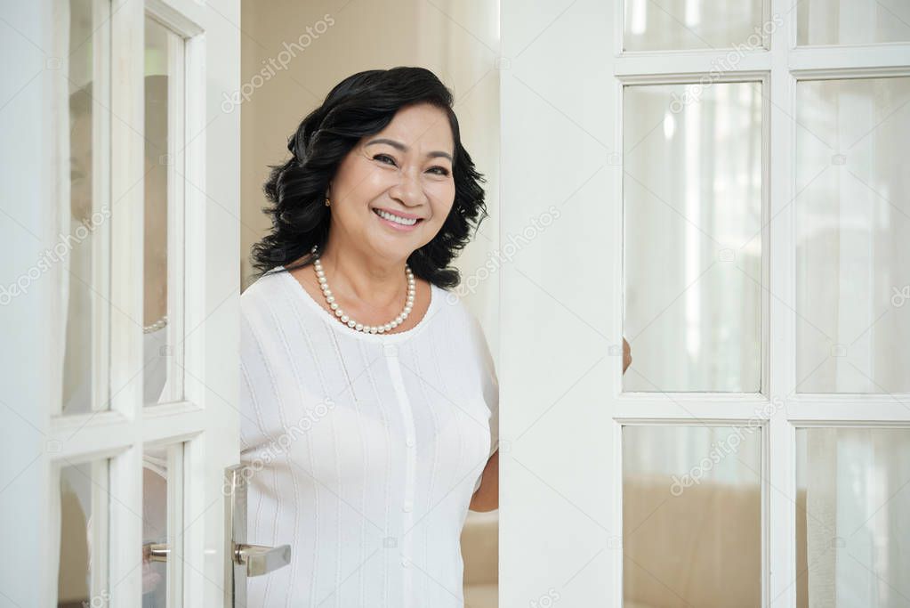 Attractive elegant mature Asian woman standing near doorway in her apartment and smiling at camera happily