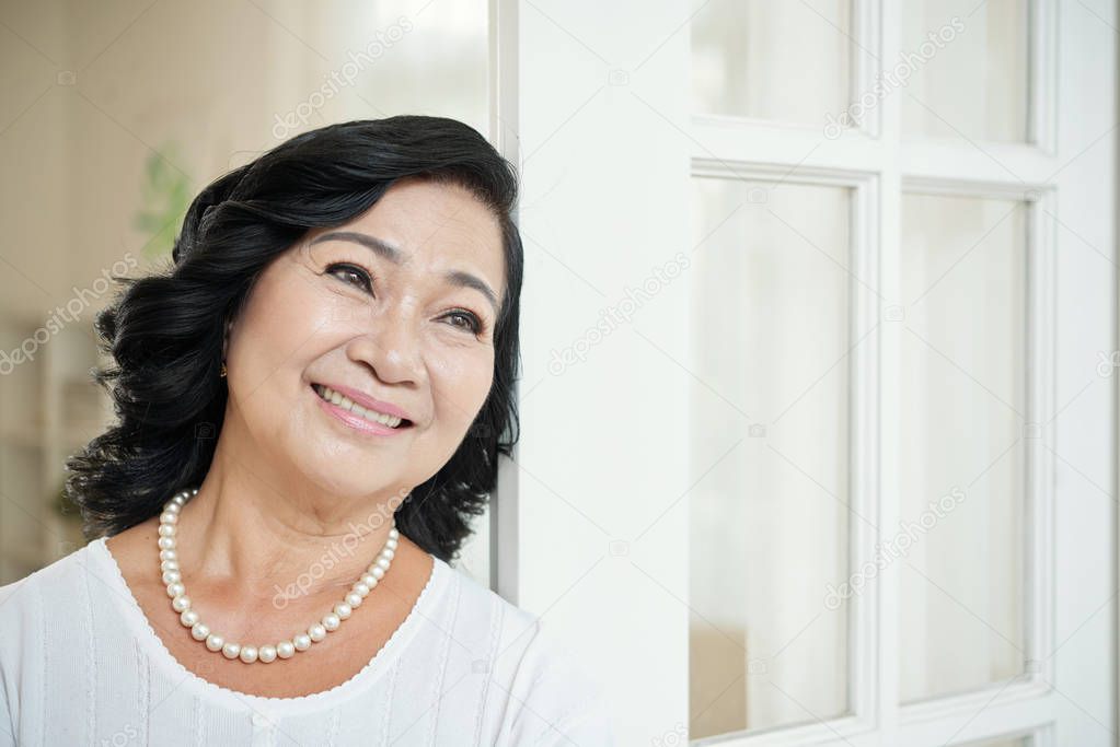 Waist-up portrait of attractive elderly Asian woman in pearl necklace leaning on doors, looking away dreamily and smiling joyfully