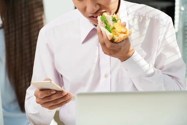 在智能手机屏幕上吃三明治和阅读信息的青年企业家 — 图库照片