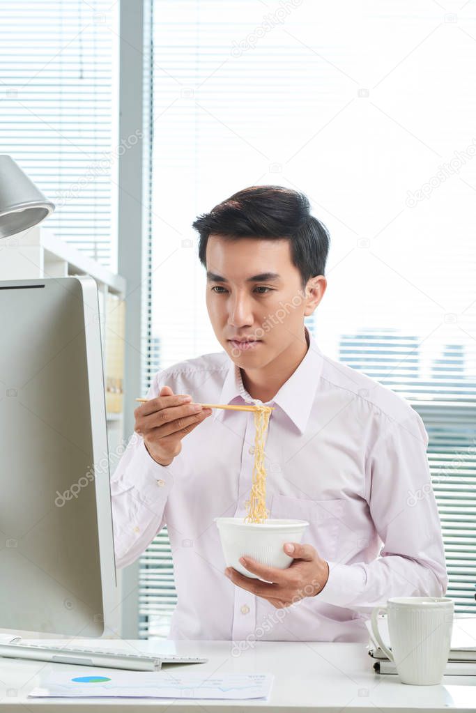 Young Vietnamese businessman reading e-mails on computer screen and eating instant noodles