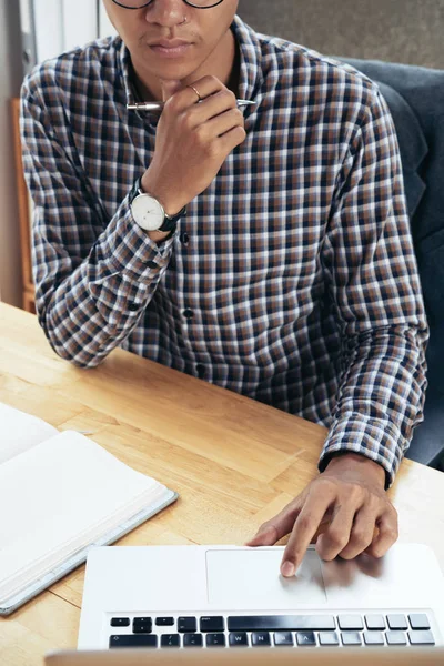 Unrecognizable young man in checked shirt sitting at table and reading information on notebook  with interest, holding chin with hand
