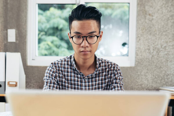 Young serious Asian man wearing checked shirt and glasses working on computer while sitting at office table