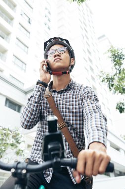 Asyalı gencin smartphone üzerinde bisiklet üzerinde otururken konuşurken gözlük ve güvenlik kask içinde düşük açılı görünüş