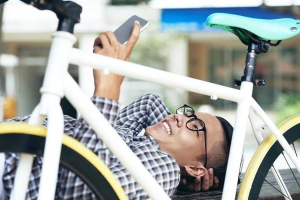 若い男性のスマート フォンでビデオ チャットを持っていることと彼の自転車の後ろに屋外に横たわっている間明るい笑顔 — ストック写真
